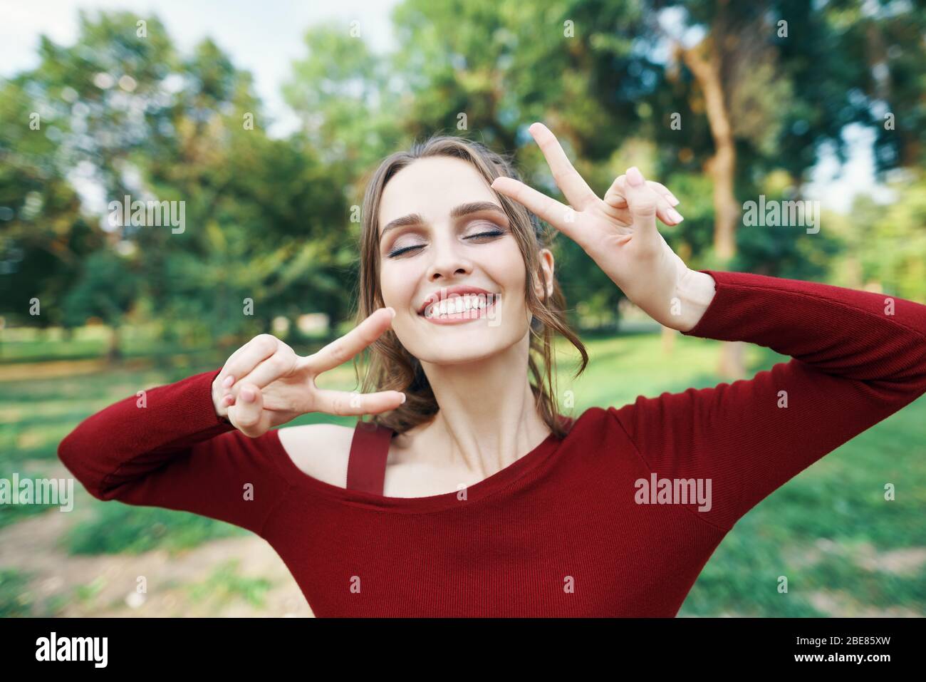 Portrait of a smiling happy woman showing victory sign posing outdoors on green summer nature background. Female beauty concept Stock Photo