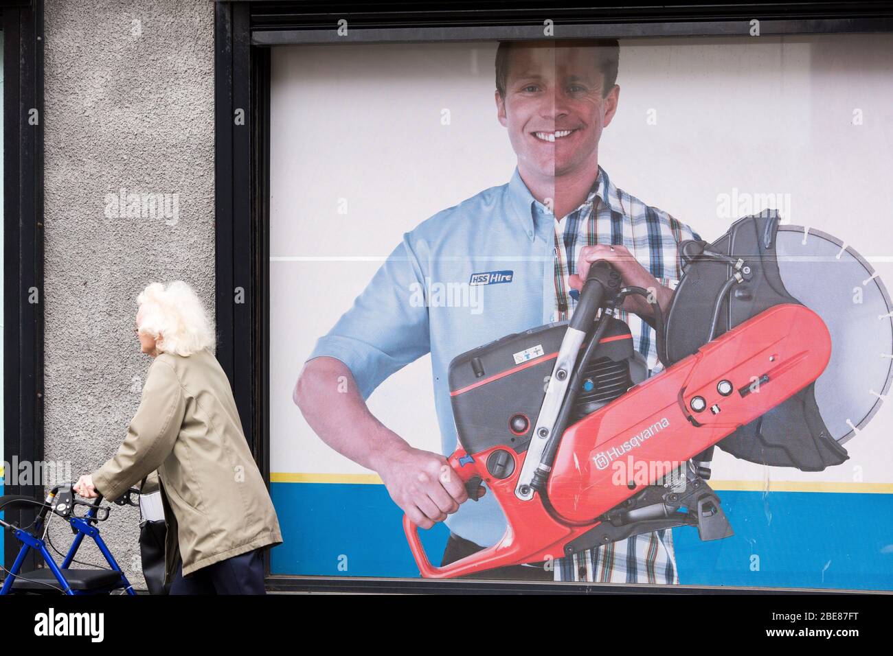 An elderly woman using a walking frame walks past a billboard for a tool hire company while walking up Picton Road in Urban Liverpool Stock Photo