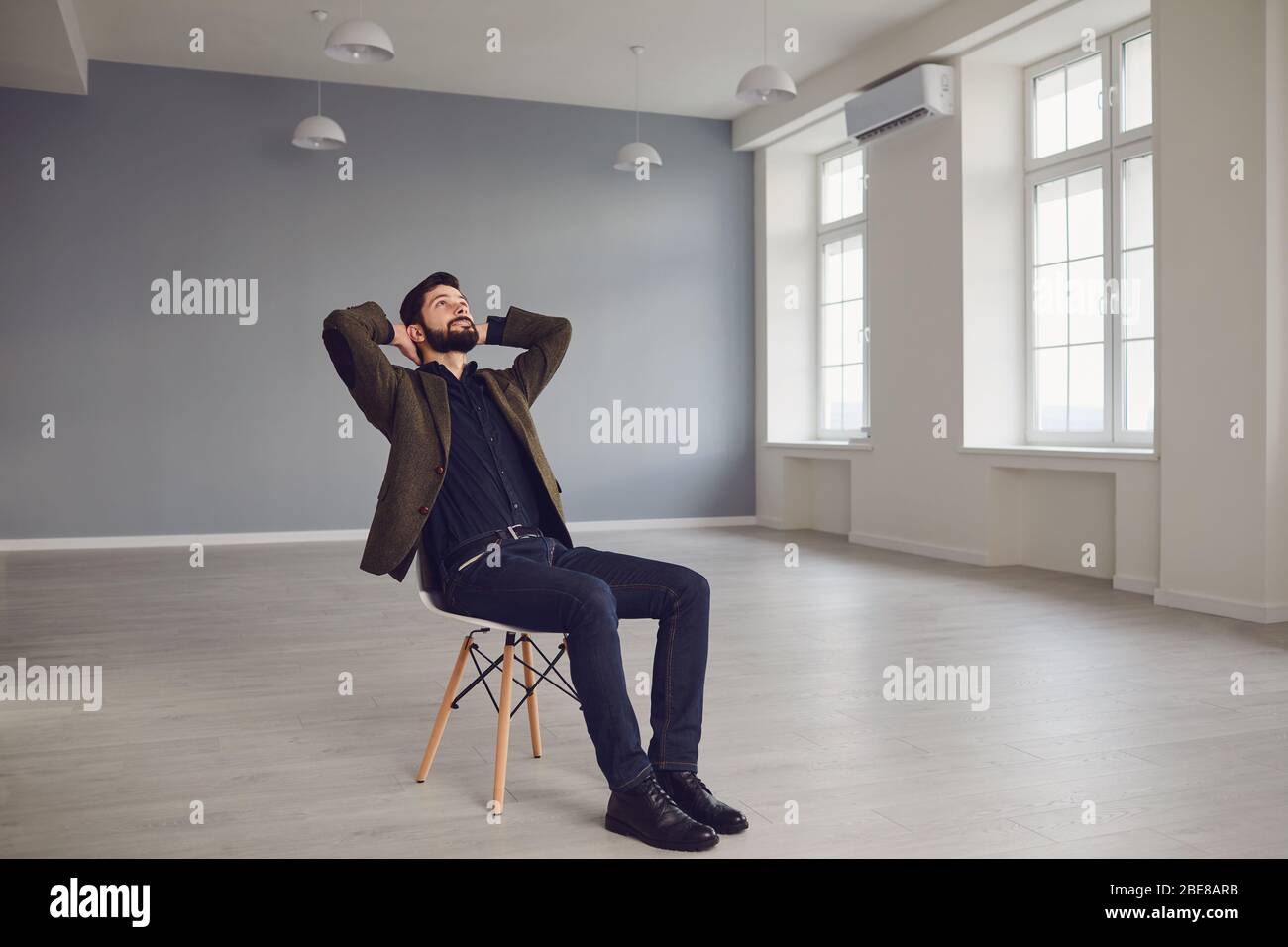 Thoughtful young male entrepreneur in elegant clothes dreaming while sitting on chair in new empty modern office room Stock Photo