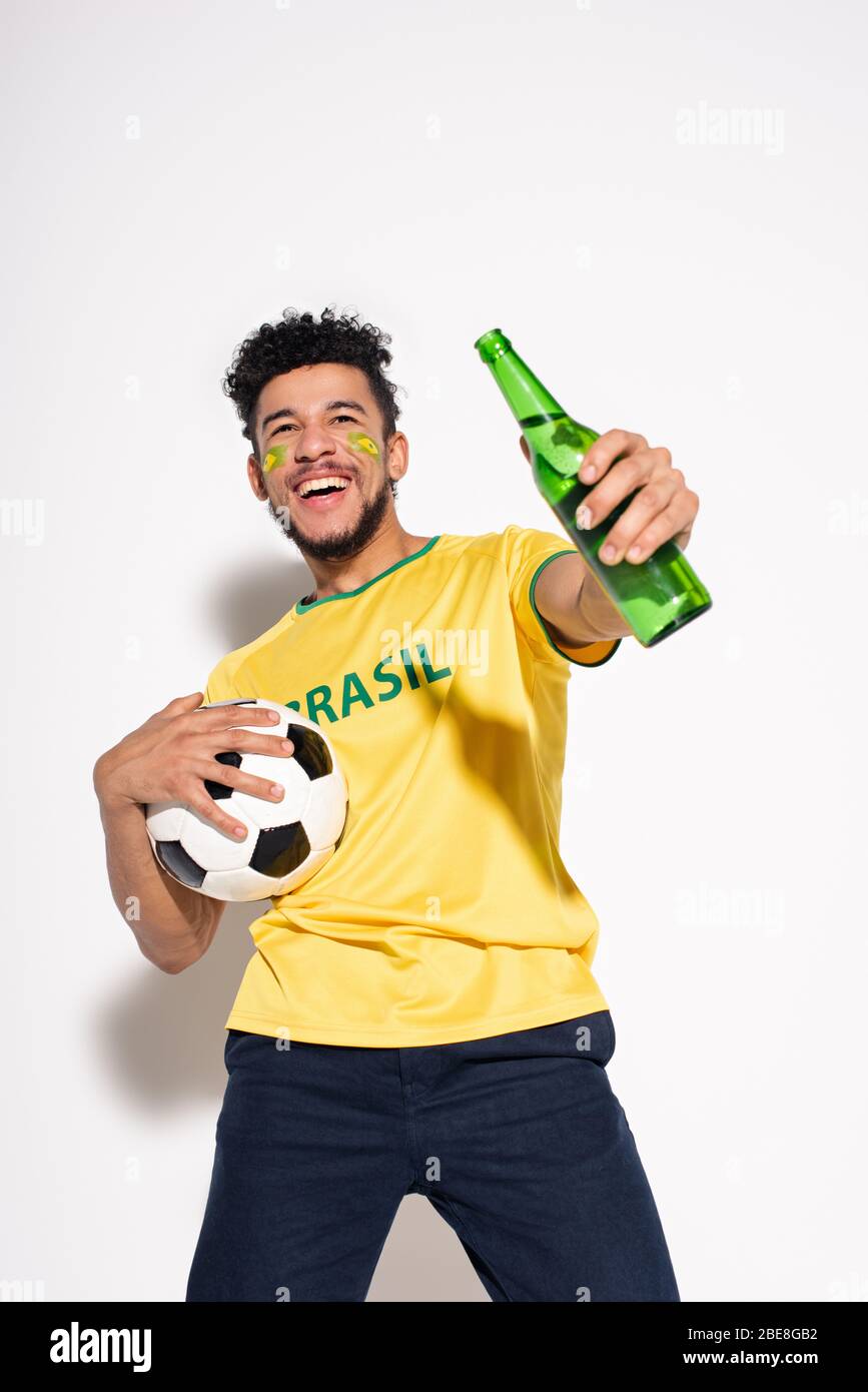 smiling african american man holding football ball and bottle of beer on grey Stock Photo