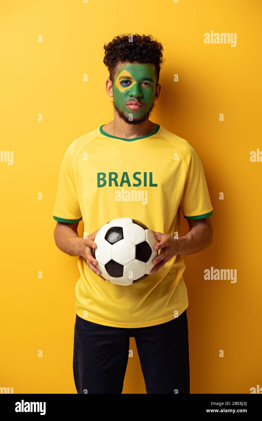 african american football fan with face painted as brazilian flag holding ball on yellow Stock Photo