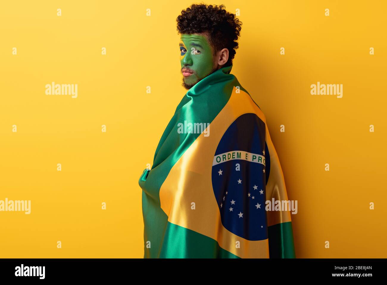 african american football fan with painted face wrapped in brazilian flag on yellow Stock Photo