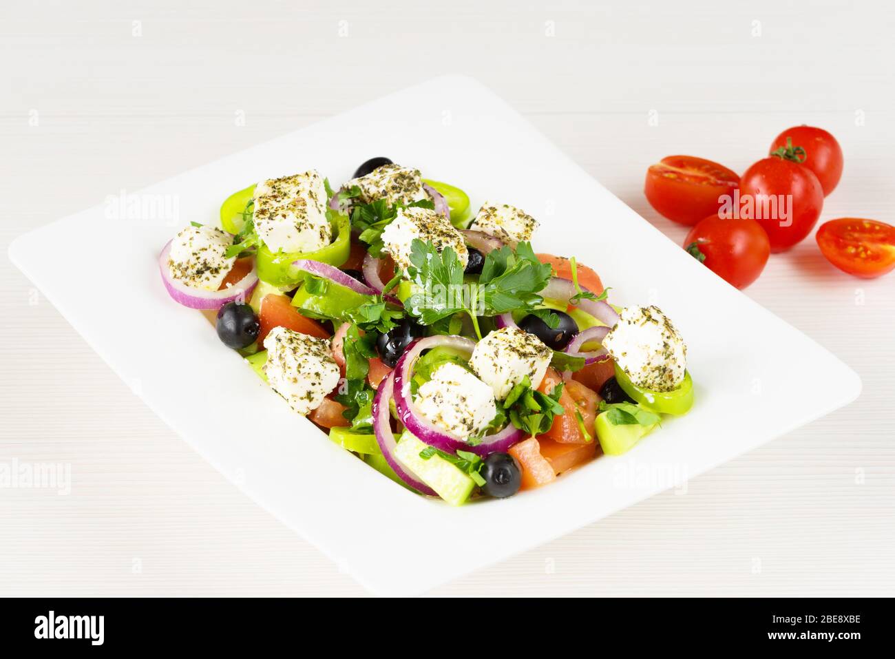 Greek salad with fresh tomato, cucumber, red onion, basil, feta cheese, black olives, Italian herbs and olive oil in white dish on white wooden table. Stock Photo