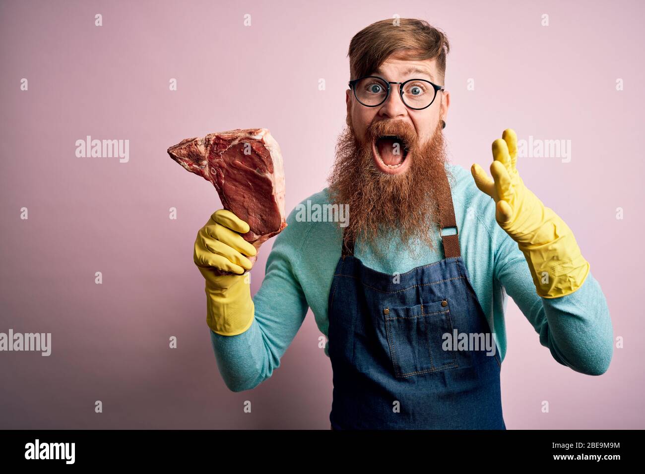 Redhead Irish butcher man with beard holding raw beef steak over pink ...