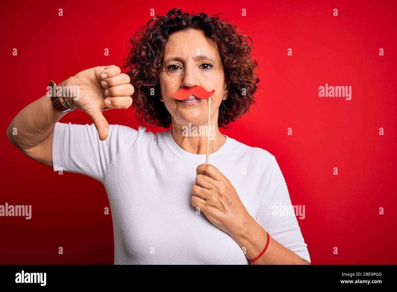 Middle age curly hair woman holding funny mustache over isolated red ...