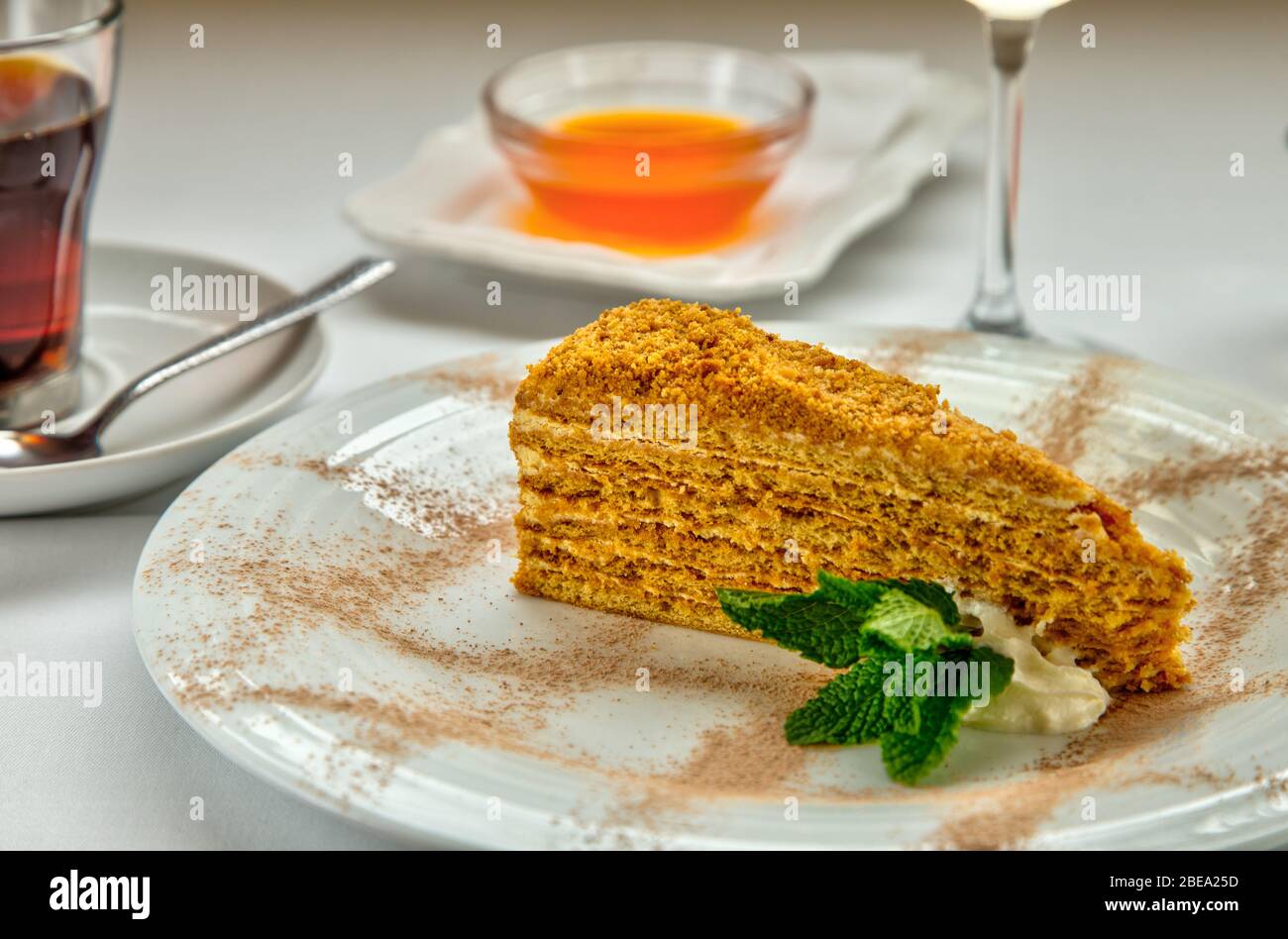 Piece of honey cake and fresh mint on white plate. Served with a rosette of honey, a cup of aromatic tea and a glass of white wine. Stock Photo