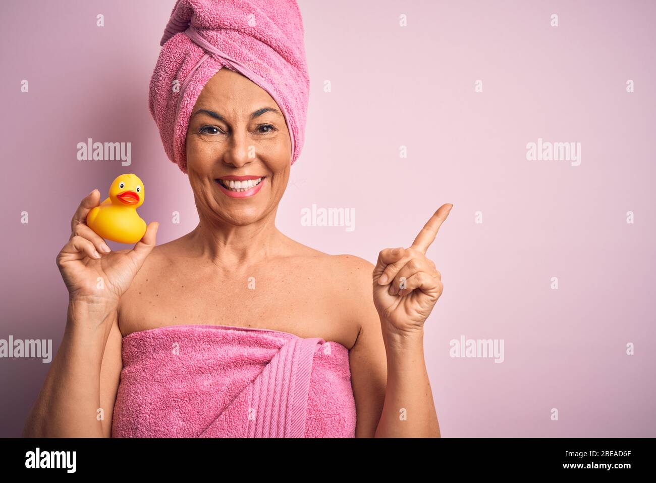 Middle age woman wearing pink bath towel from beauty body care holding rubber yellow duck very happy pointing with hand and finger to the side Stock Photo