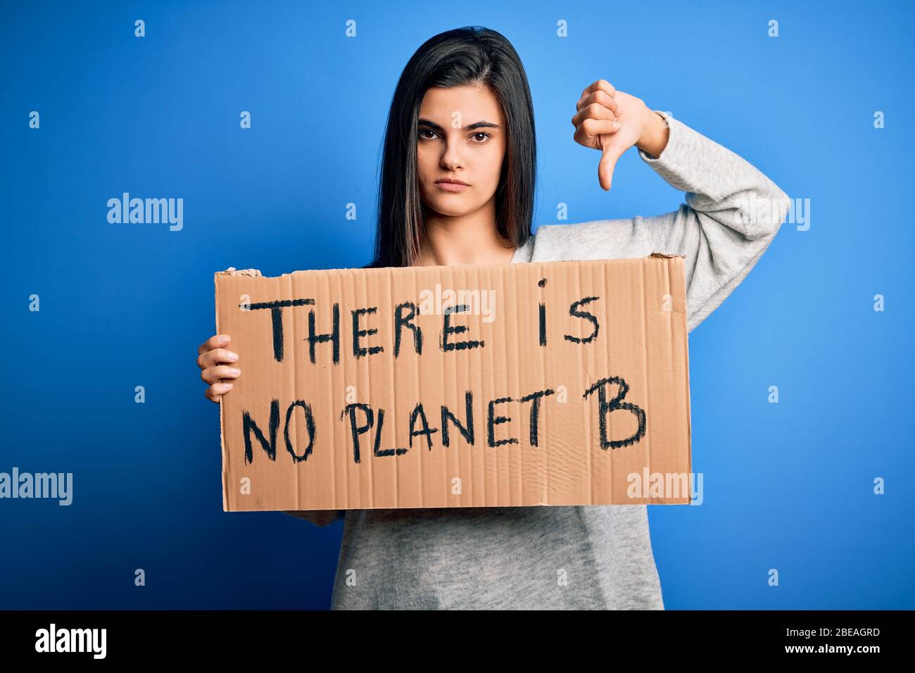 Young beautiful brunette activist woman holding banner protesting to care the planet with angry face, negative sign showing dislike with thumbs down, Stock Photo