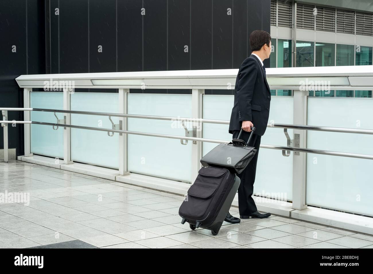 A businessman dressed in a black suit with a rolling suitcase (carry-on) and a briefcase. Stock Photo