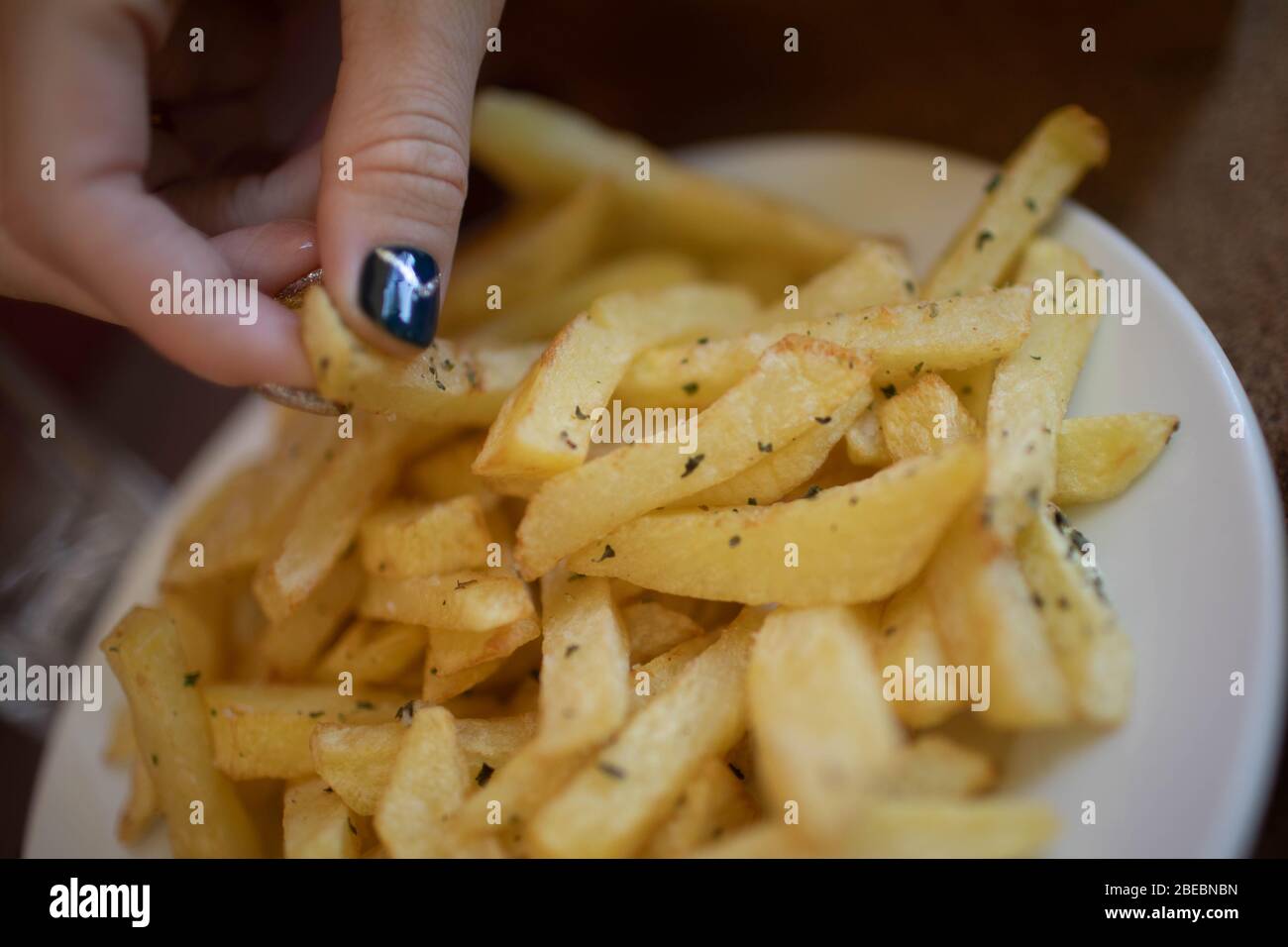 French fries on a white plate. A woman's hand reaches for a potato. Stock Photo