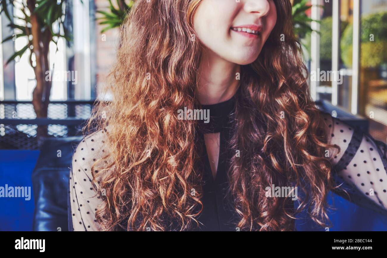 A young lady with curly brown hair rests in the cafeteria, leaning on the back of the chair and looking away. We only see the lower half of her face that looks away from the camera with a light smile on her lips. Toned image. Stock Photo