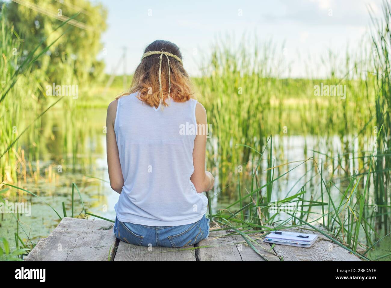 Young woman sitting with her back on wooden pier, enjoying picturesque view of lake water, relaxing in nature on sunny summer day, copy space Stock Photo