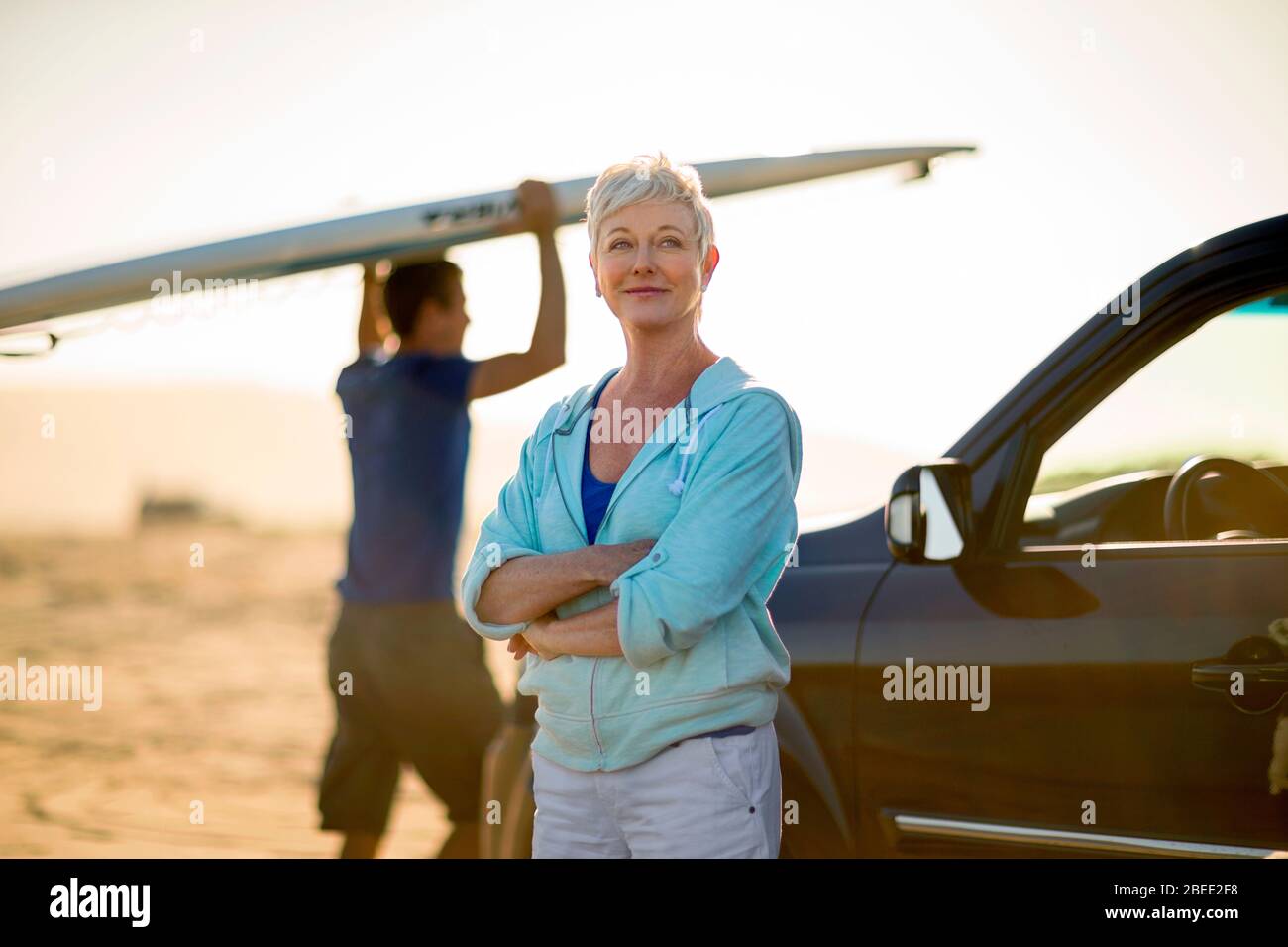 Portrait of a content mature woman standing by her car while a mid-adult man walks toward beach with a surfboard Stock Photo