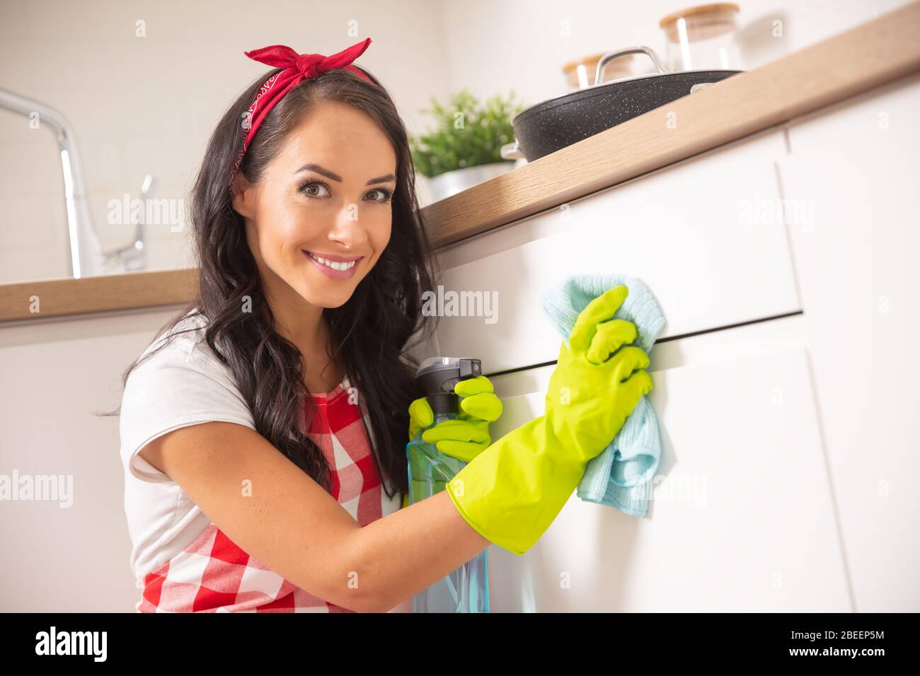 Lovely young housemaid with red ribbon in her hair and apron cleaning white kitchen cupboard door with a cloth and detergent in yellow rubber gloves. Stock Photo