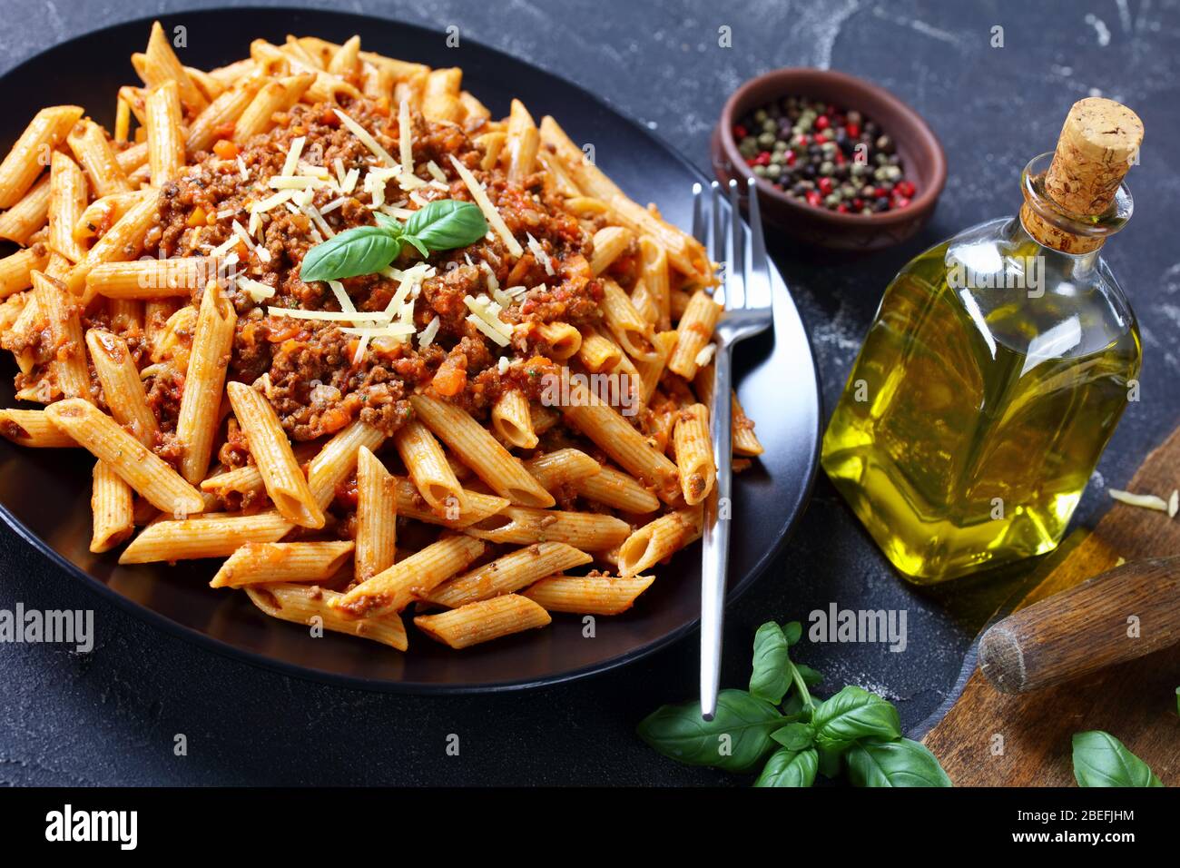 wholewheat pasta penne bolognese topped with shredded parmesan and basil on a black plate on a concrete table with ingredients, horizontal view from a Stock Photo