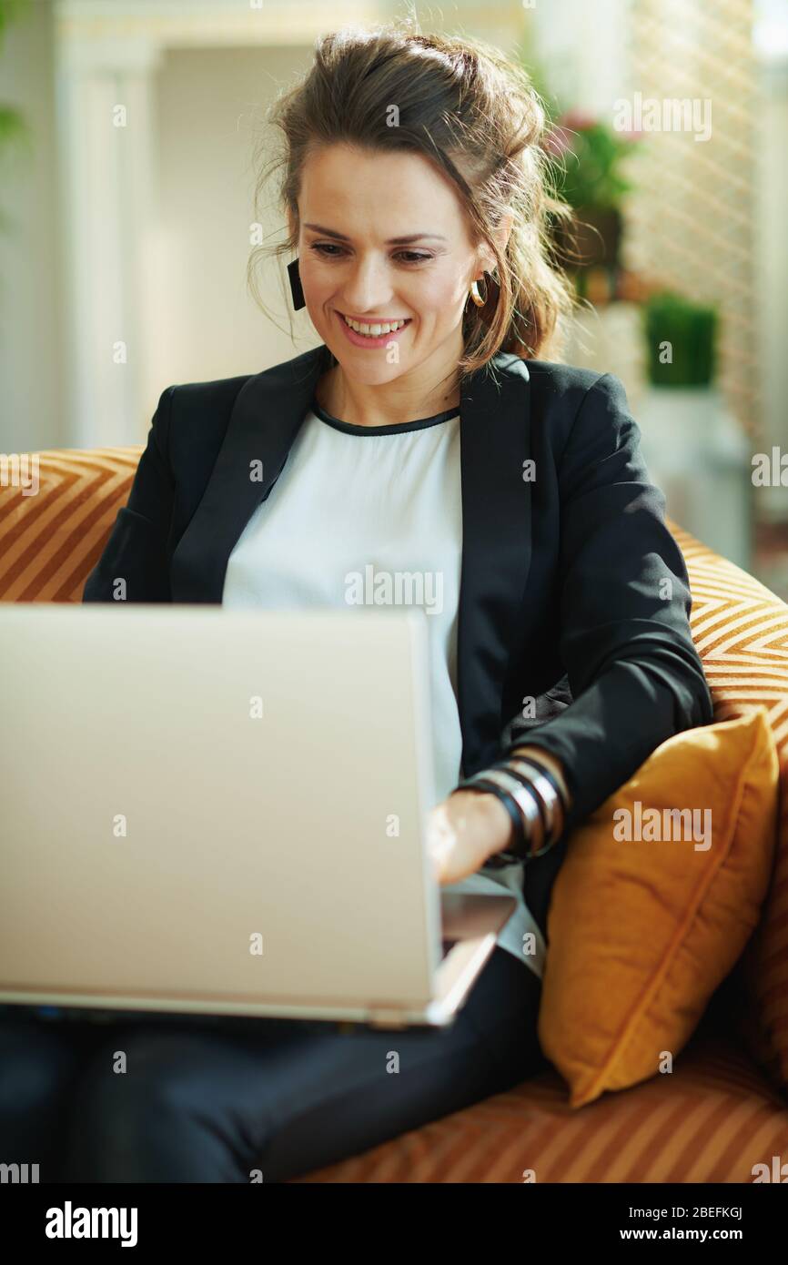smiling stylish woman in white blouse and black jacket at modern home in sunny day doing research on a laptop while sitting on divan. Stock Photo