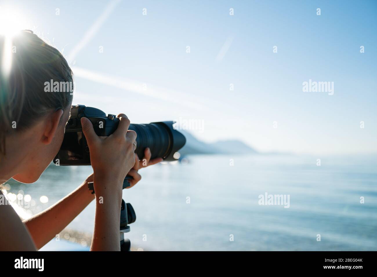 Professional female photographer taking photo of beautiful morning sea with her camera on tripod, with rising sun flare shining above her head. Stock Photo