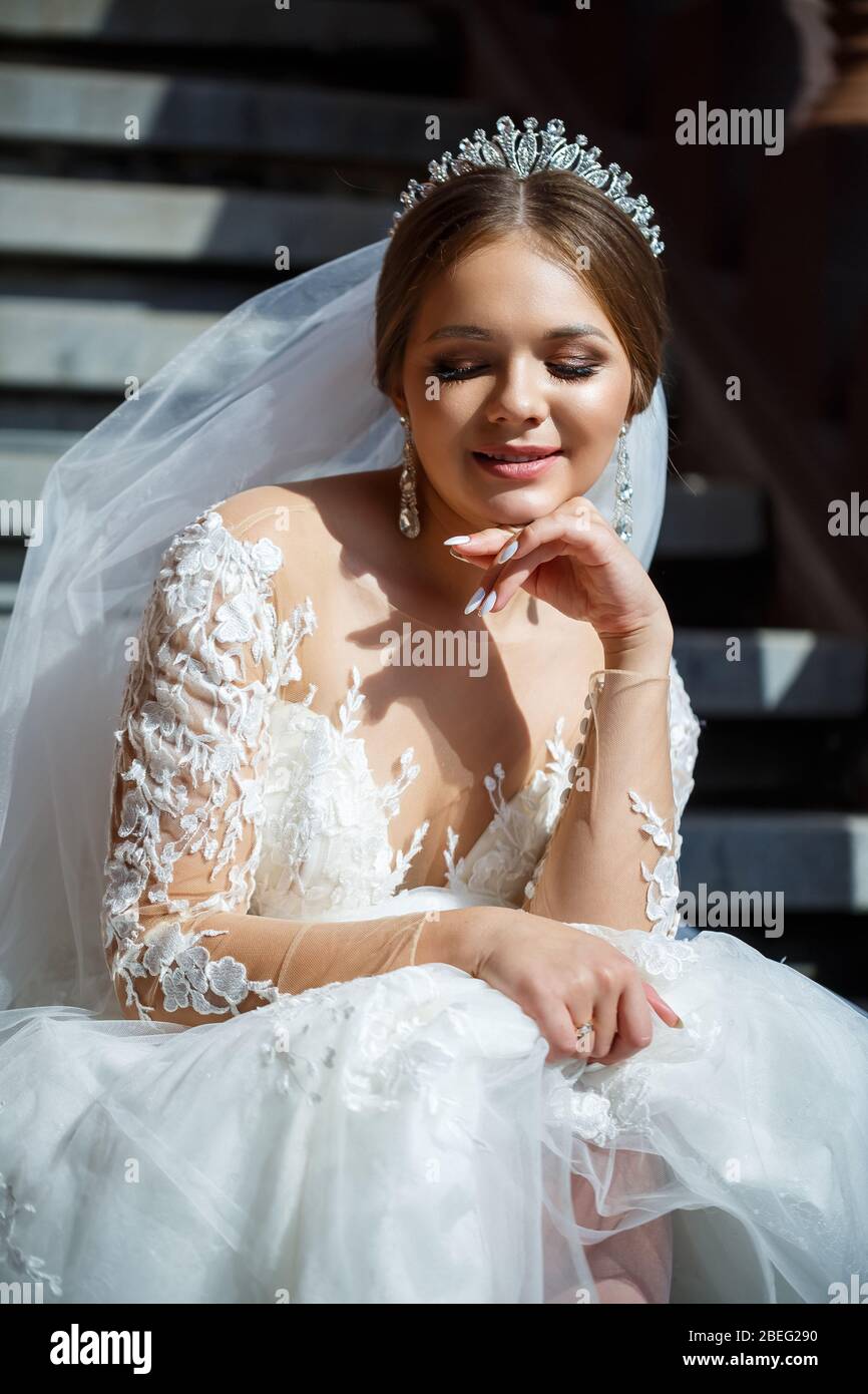 Beautiful bride in a white wedding dress sits on the steps and smiles ...