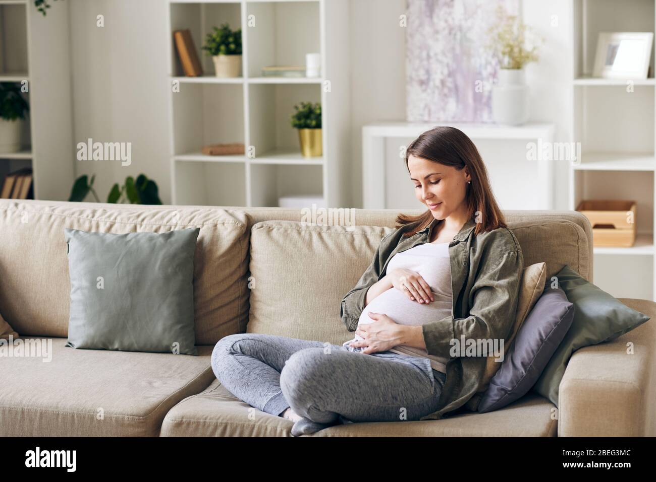 Calm attractive young pregnant woman sitting with crossed legs on sofa
