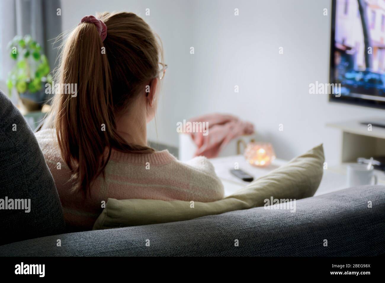 A young woman from behind with ponytail and glasses watching TV while sitting on sofa in living room at home. Binge watching tv via online streaming Stock Photo