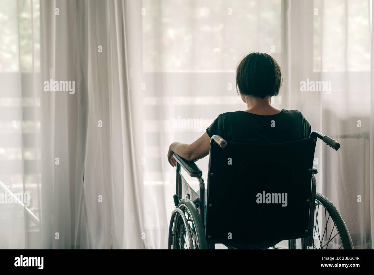 Depressed sad woman in worn wheelchair looking out the window and thinking, lonely disabled adult caucasian person indoors Stock Photo