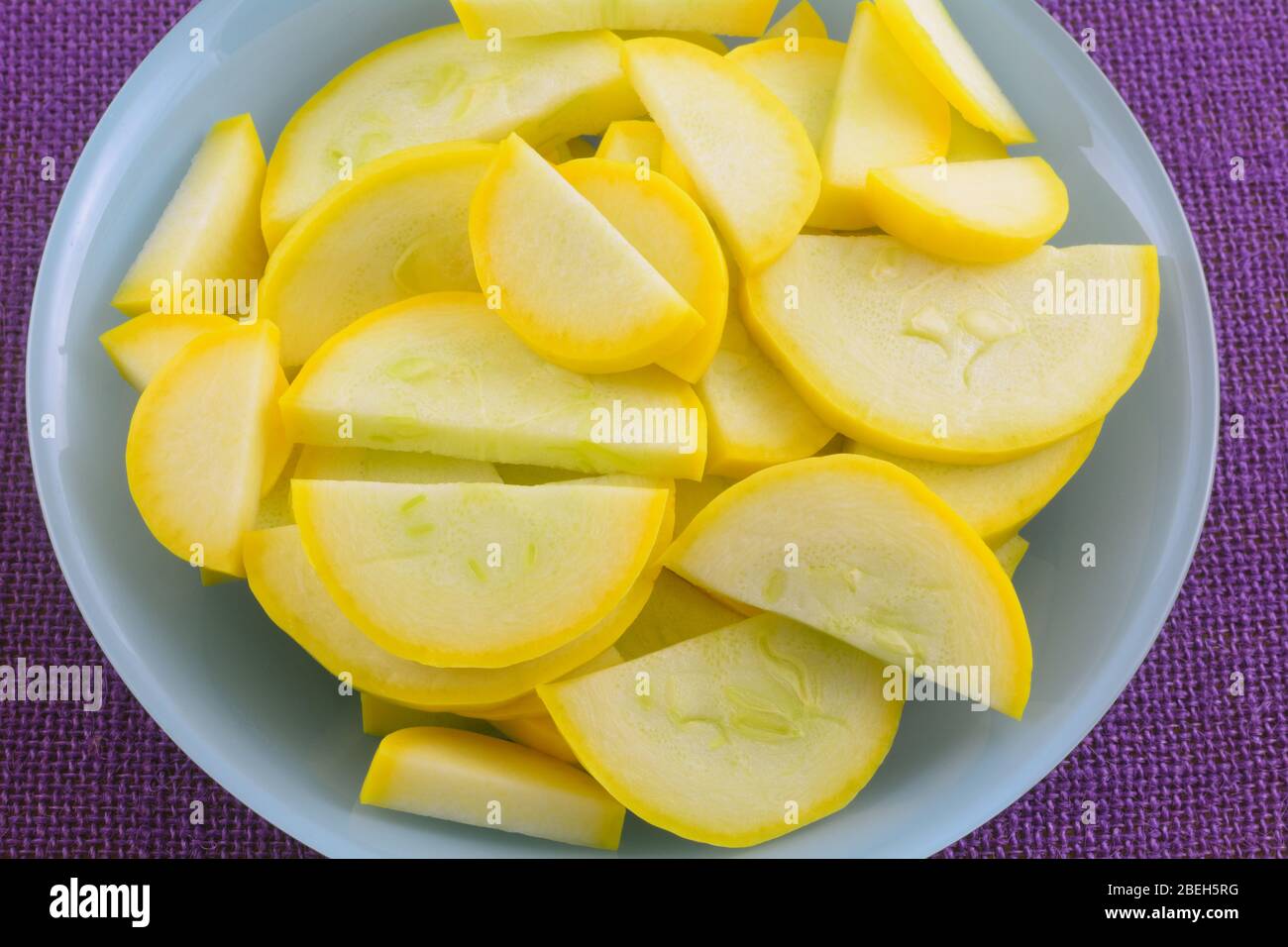 Sliced raw yellow summer crookneck squash in blue bowl on purple