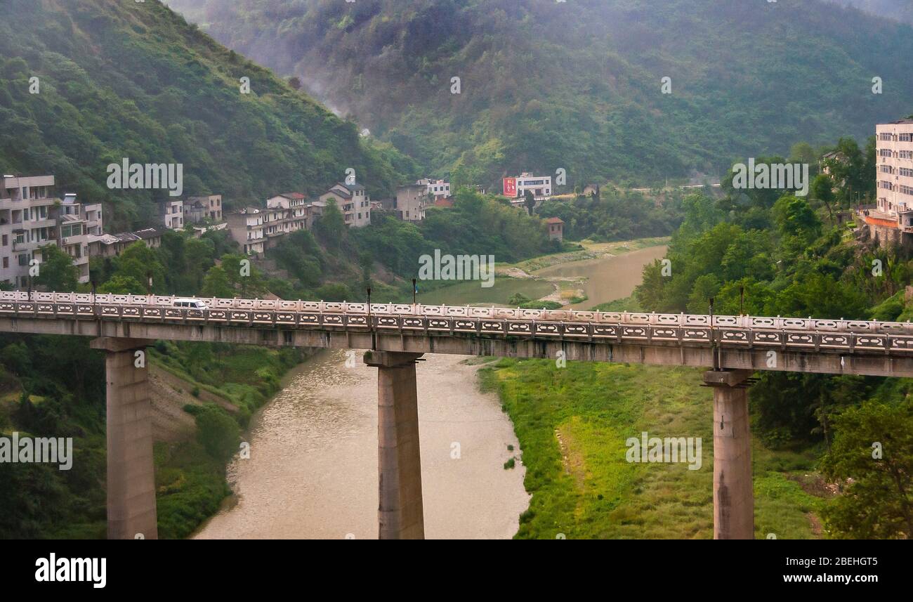 Yichang, China - May 5, 2010: Bridge on pillars over brown meandering rivers between green mountain slopes with houses along a road. Stock Photo