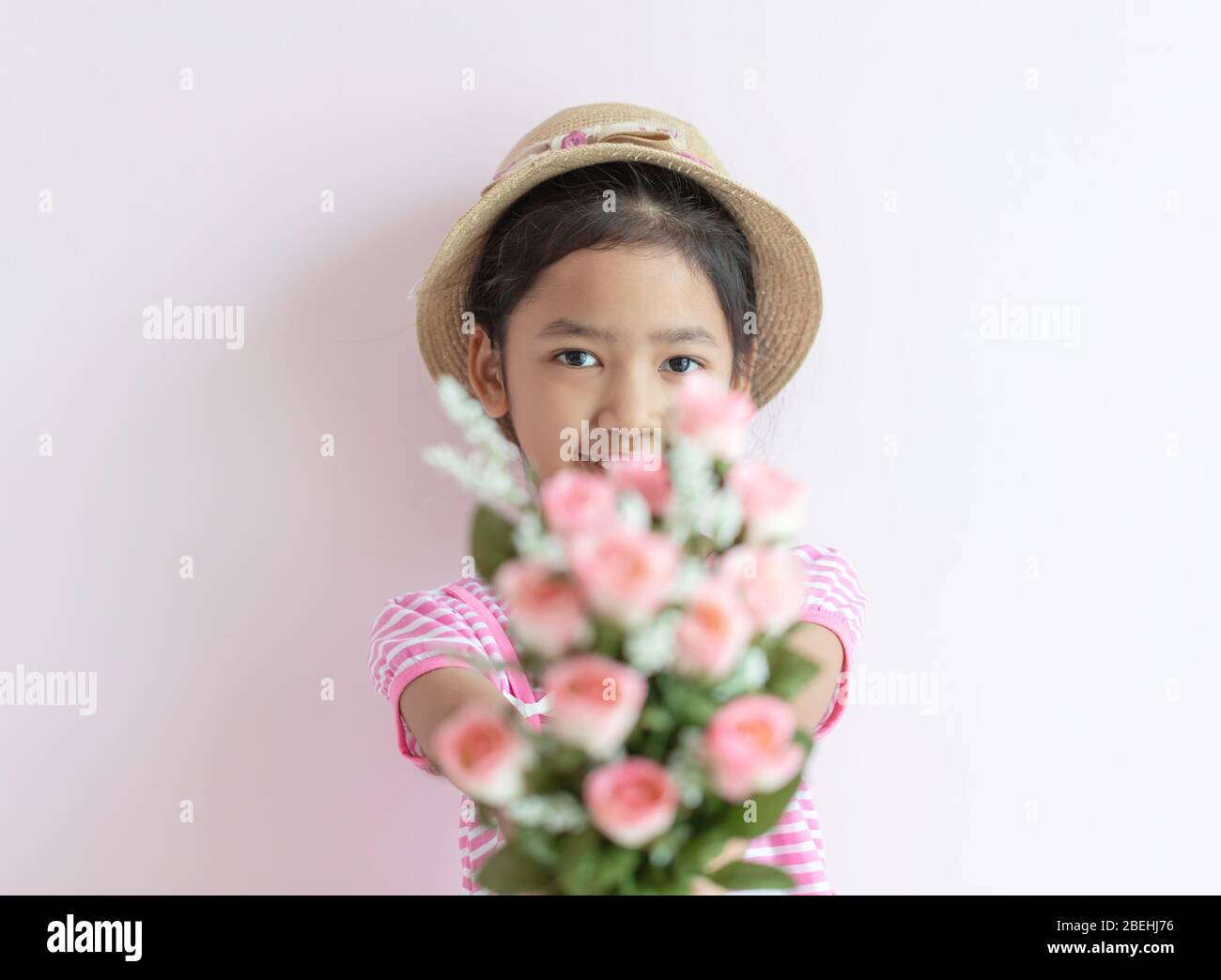 Portrait of an Asian little girl wearing a pink and white striped dress. The child wears a hat and holding roses flowers with smiling and happy. Selec Stock Photo