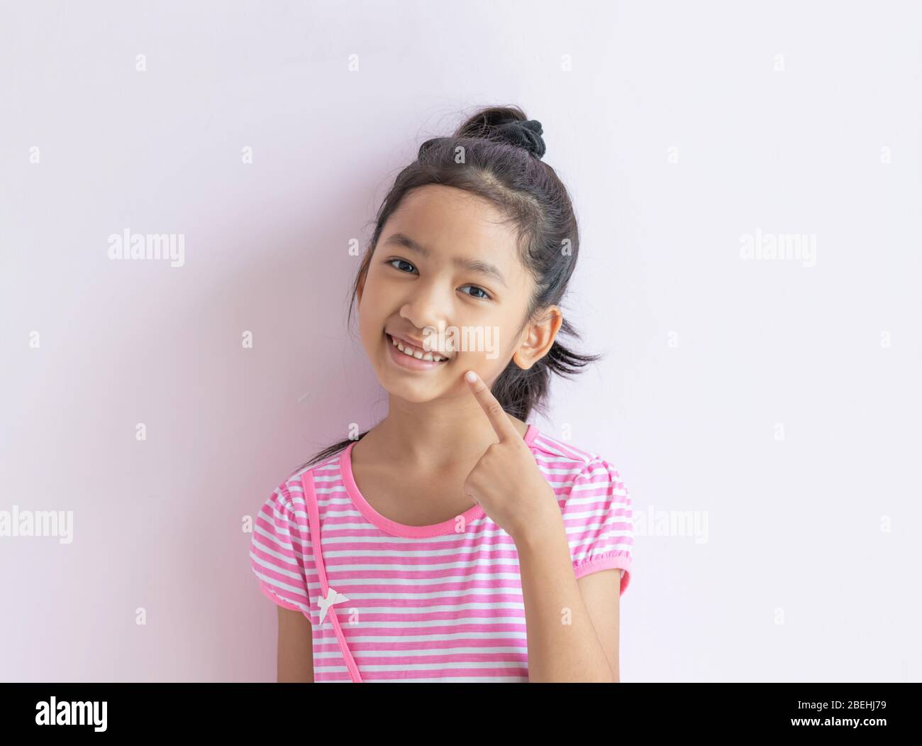 Portrait of an Asian little girl wearing a pink and white striped dress. The child with black hair is smiling and pointing to her face. Stock Photo