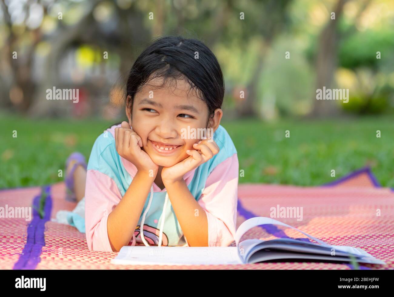 The Asian little girl lay down and put her hand under the chin on the book with a smile on the lawn with sunray in the garden. The kid reading a book Stock Photo