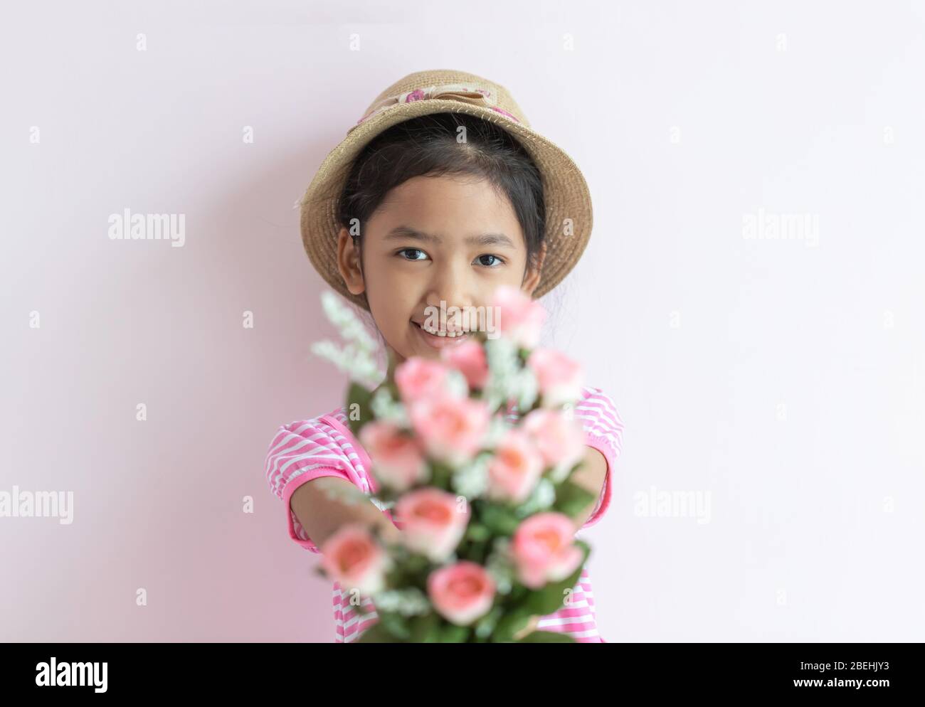 Portrait of an Asian little girl wearing a pink and white striped dress. The child wears a hat and holding rose flowers with smiling and happy. Select Stock Photo