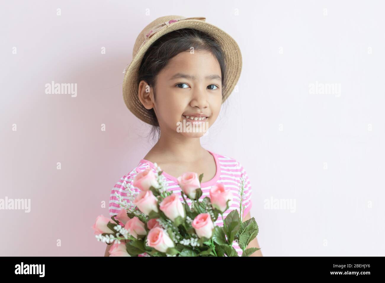 Portrait of an Asian little girl wearing a pink and white striped dress. The child wears a hat and holding roses flowers with smiling and happy. Stock Photo