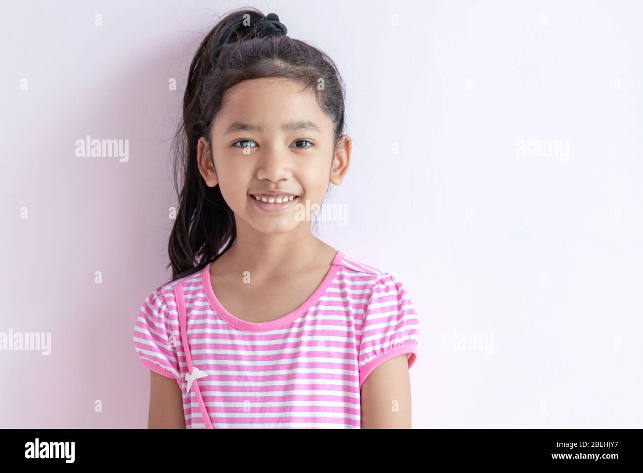 Portrait of an Asian little girl wearing a pink and white striped dress. The child with black hair is smiling brightly. Stock Photo