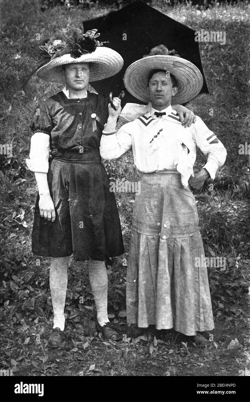 Two men, both dressed head to toe in women's clothing (except their shoes), c.1906. For the Edwardian era, both are finely dressed in casual summertime clothing, including hugely wide-brimmed, flowered hats. The man holding the open umbrella (while standing in the shade) had a dainty handkerchief pinned to his white blouse.  Are they a loving couple? friends? Gay?  Perhaps they are dressed in costumes for a play.   To see all my related images Search:  Prestor  vintage  gay Stock Photo
