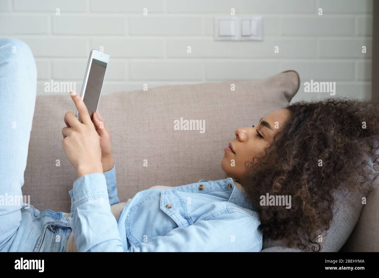 Young african woman using digital tablet relaxing lying on sofa at home. Stock Photo