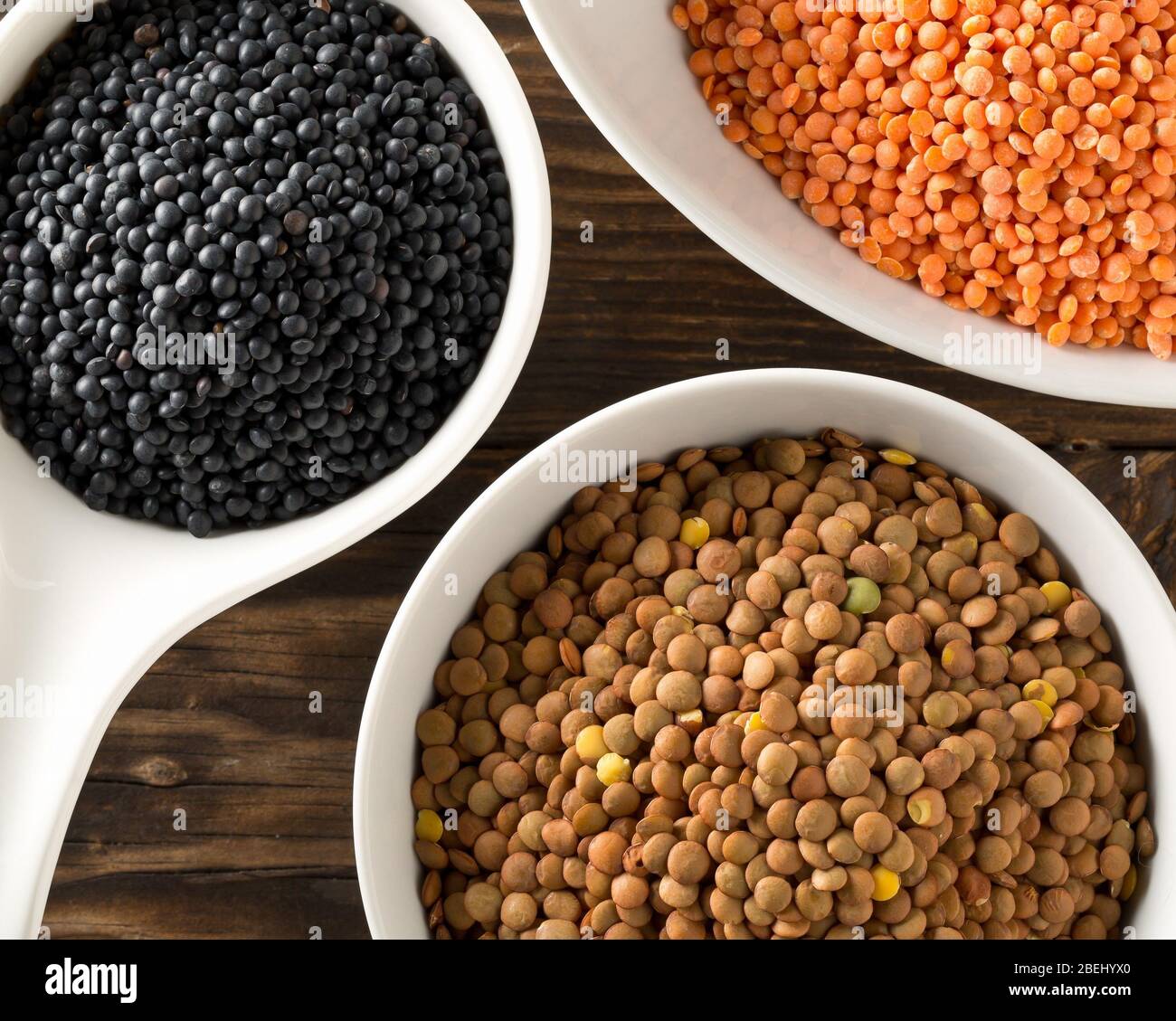 Different assorted lentils mix with red, brown and black beluga lentils in white bowls on brown wooden table background top view flat lay from above - Stock Photo