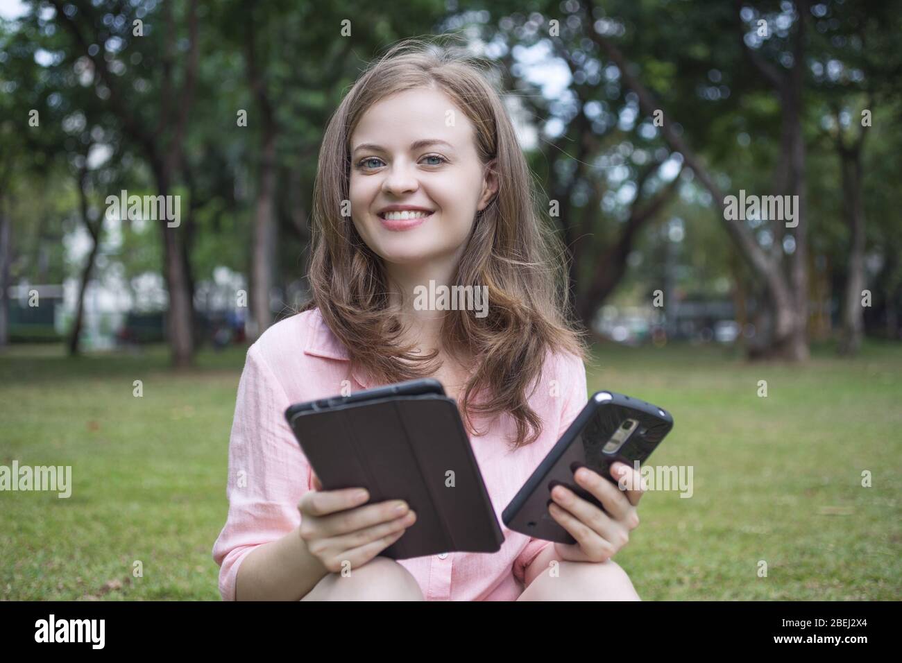 Young caucasian woman with smartphone in one hand and tablet in another looking at the camera and smiling. Outdoor portrait. Stock Photo