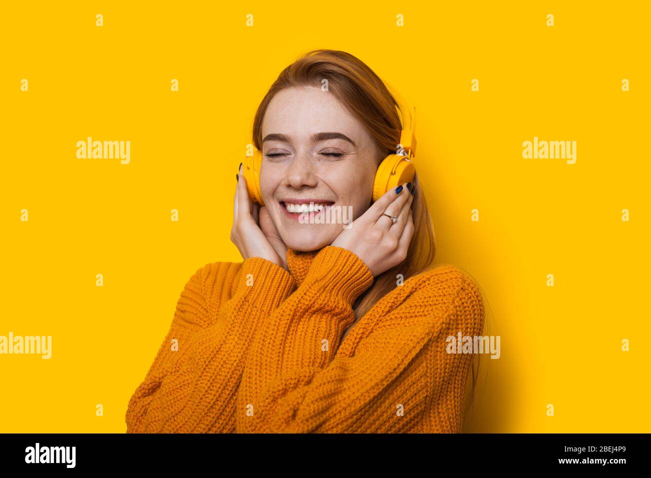 Close up photo of a ginger girl with freckles listening to music through headphones and smiling cheerfully in a sweater on a yellow wall Stock Photo