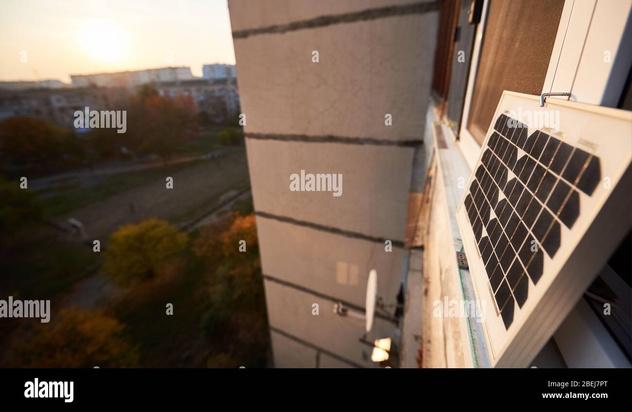 Solar panel on the window of a high-rise residential building against a background of a sleeping area of the city with clear blue sky on a warm day. Converting solar radiation into electricity Stock Photo