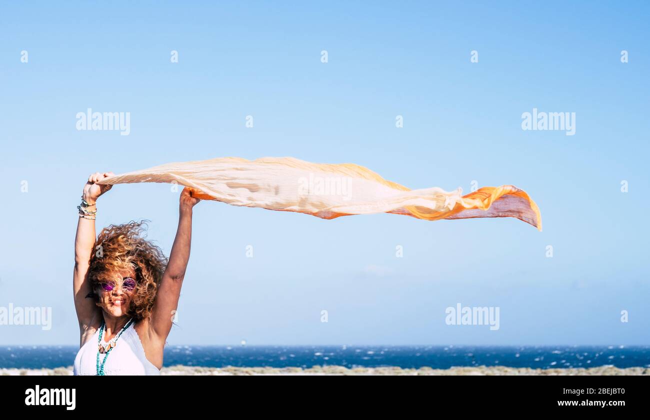 Happy and cheerful caucasian young woman play with the wind and orange textile with blue sky and ocean background - concept of freedom and happiness f Stock Photo