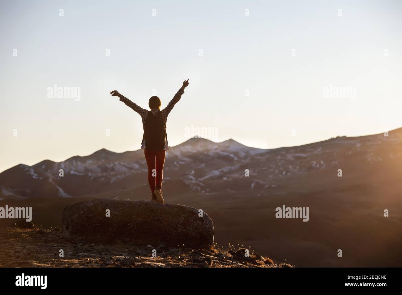 Happy female hiker with backpack stands with raised arms on big rock and looks on mountains and sunset Stock Photo