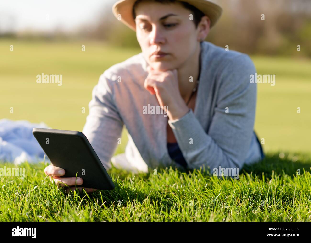 Woman in grey top, straw hat  looking at camera and smiling,  holding ebook in her hands, lying and resting on meadow / green grass on sunny spring wa Stock Photo