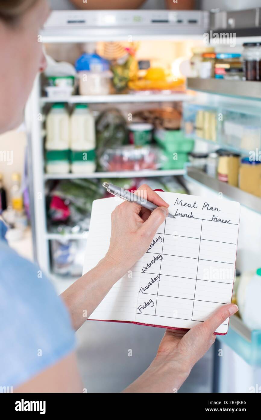 Woman Standing In Front Of Refrigerator In Kitchen With Notebook Writing Weekly Meal Plan Stock Photo