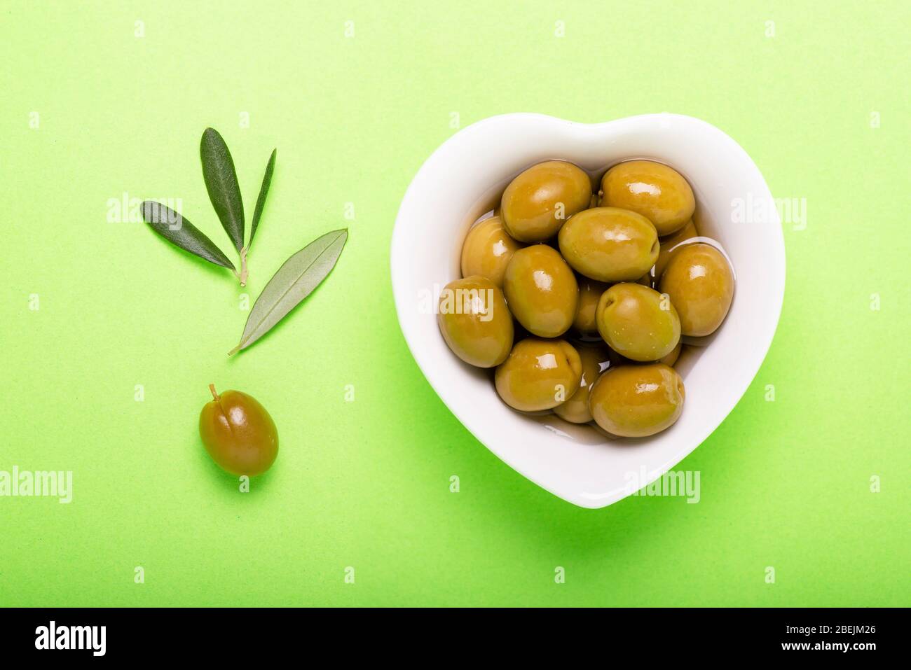 top view, on the green uniform background, white ceramic heart-shaped bowl with pickled olives Stock Photo