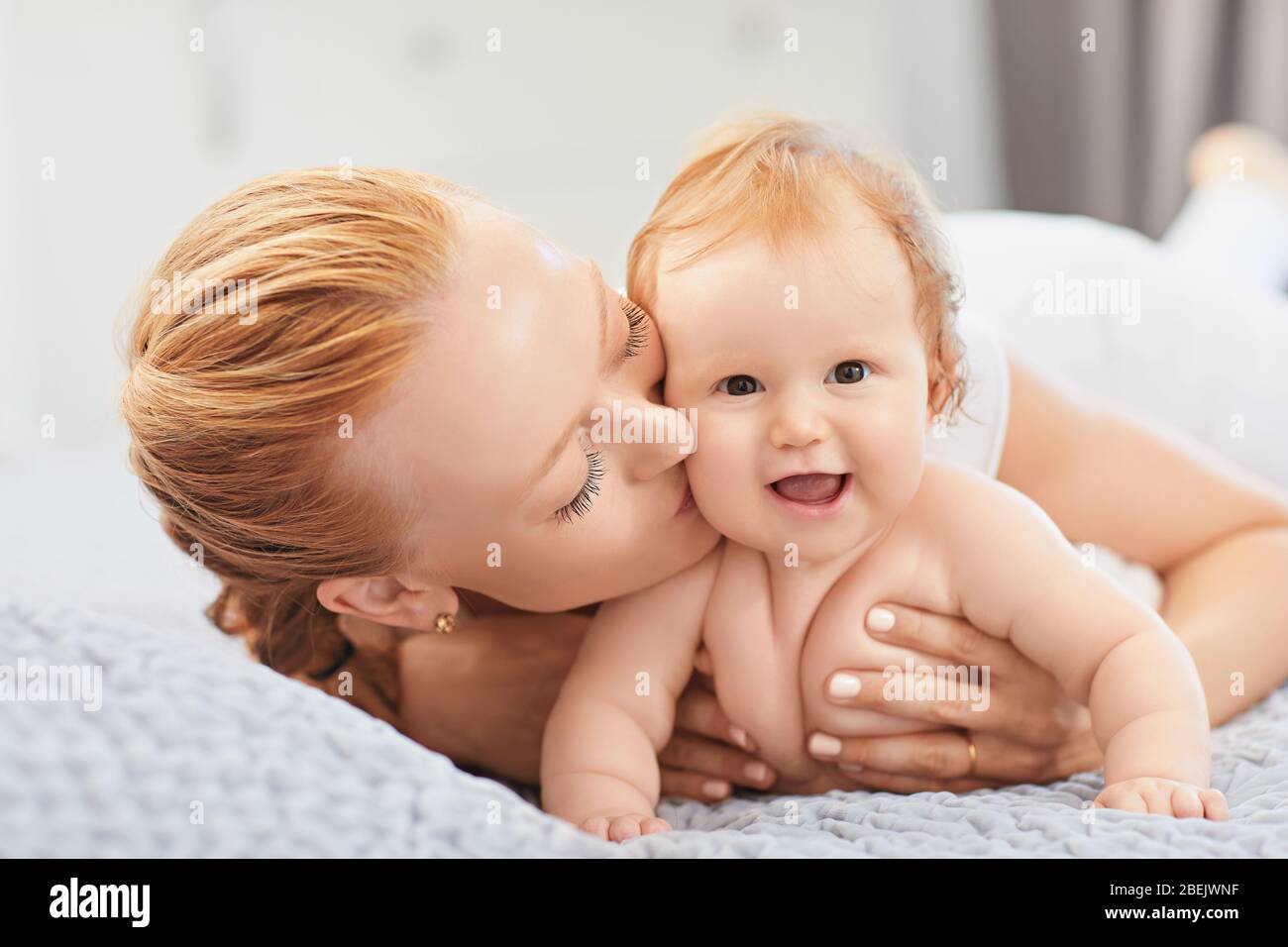 Happy mother smiles at baby lying on bed in room. Stock Photo