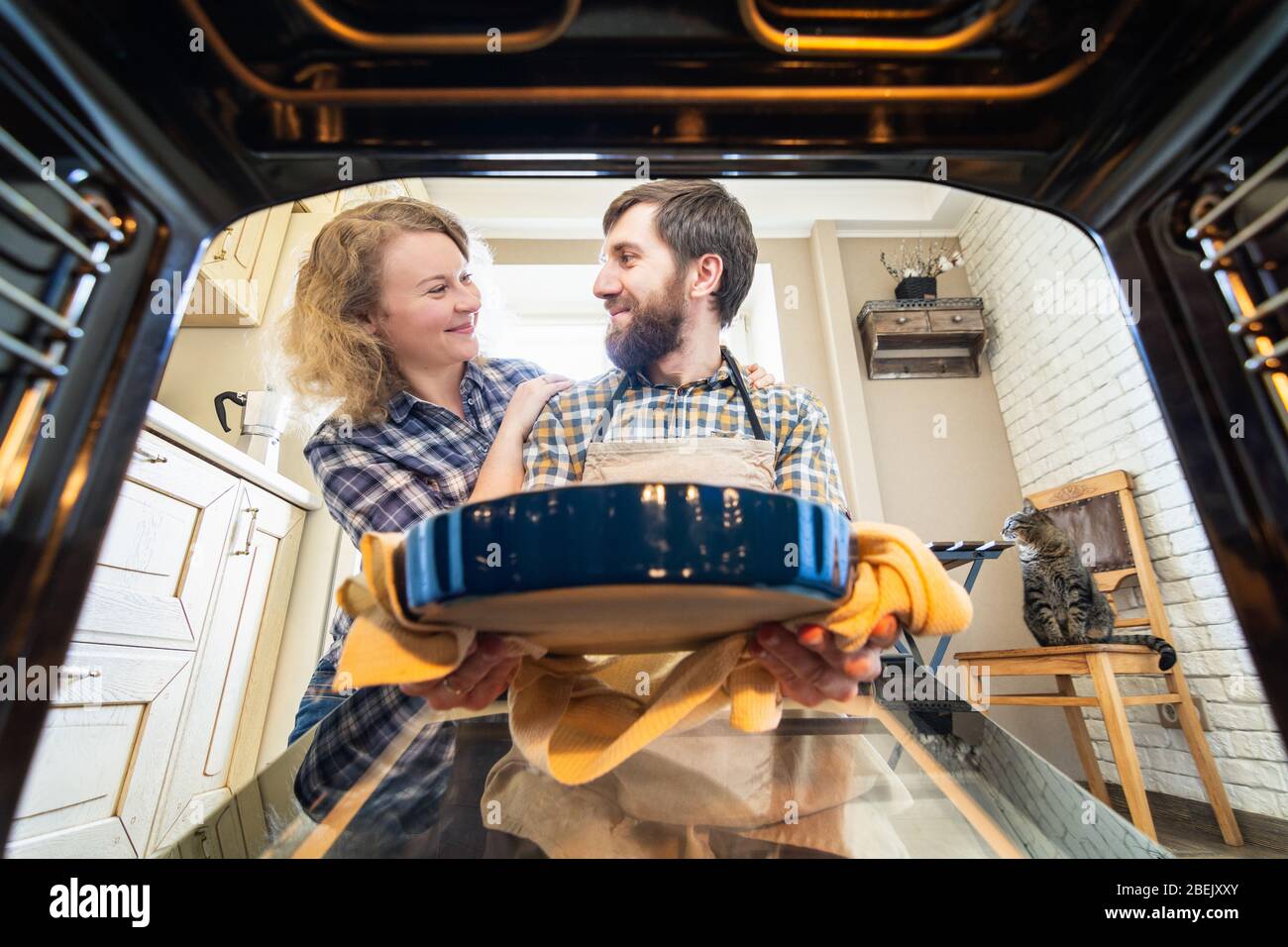 Eastern European couple cooking at home, smiling and looking at each other. Man taking out a pie from the oven with kitchen interior and a cat on back Stock Photo