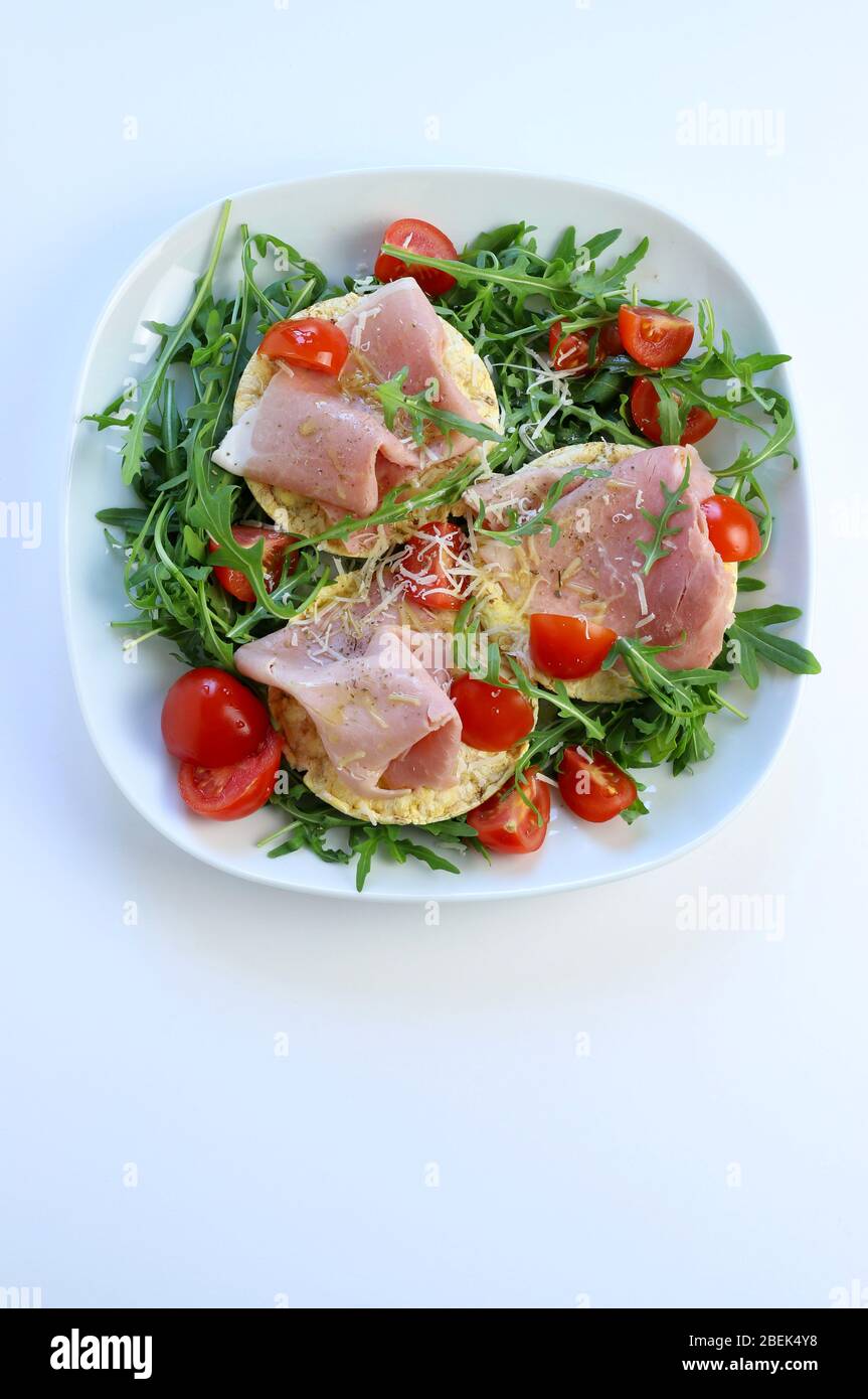 Concept of healthy food. Bio organic corn cakes with ham, fresh rocket salad and tomatoes on a plate, white background. Top view. Stock Photo