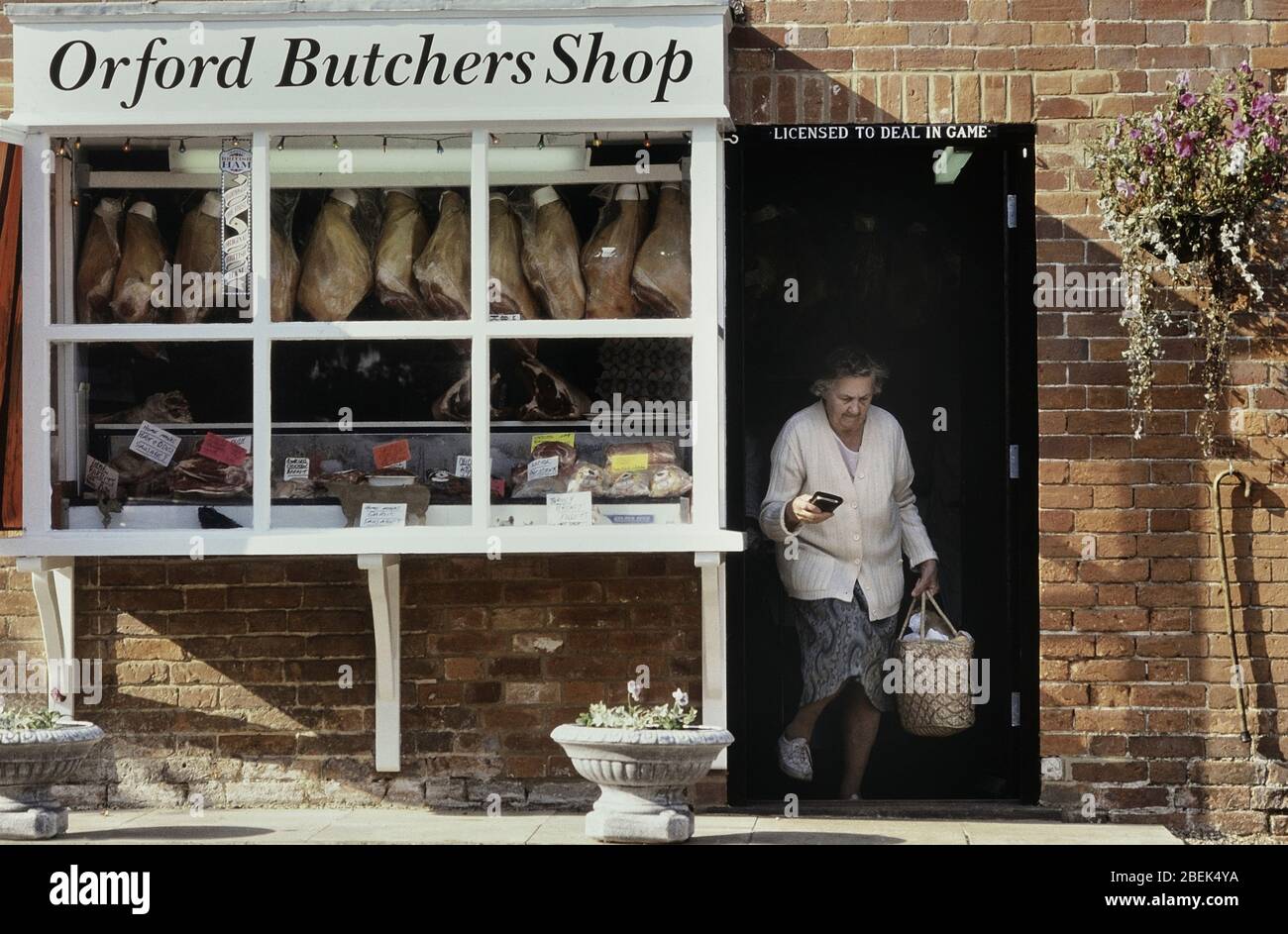A mature woman carrying a shopping basket leaving a traditional local butchers shop in Orford. Woodbridge, Suffolk, England. UK Stock Photo