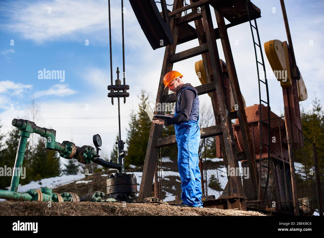 Male engineer, wearing blue overalls and orange helmet, with a laptop checking work of pump jack, making notes in his computer. Concept of petroleum industry and oil extraction. Stock Photo