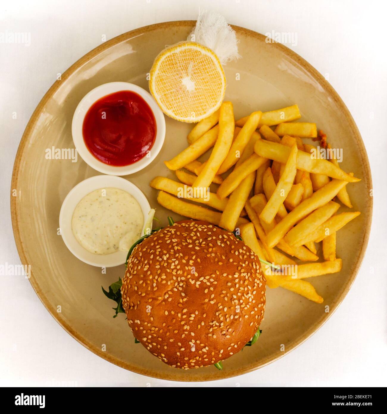Flat lay of a burger and chips / French fries served with tomato ketchup, tartar sauce, and lemon on the side on white background Stock Photo
